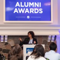 Suzanne Zack reaching out hand as she talks with 2024 Alumni Awards sign behind her